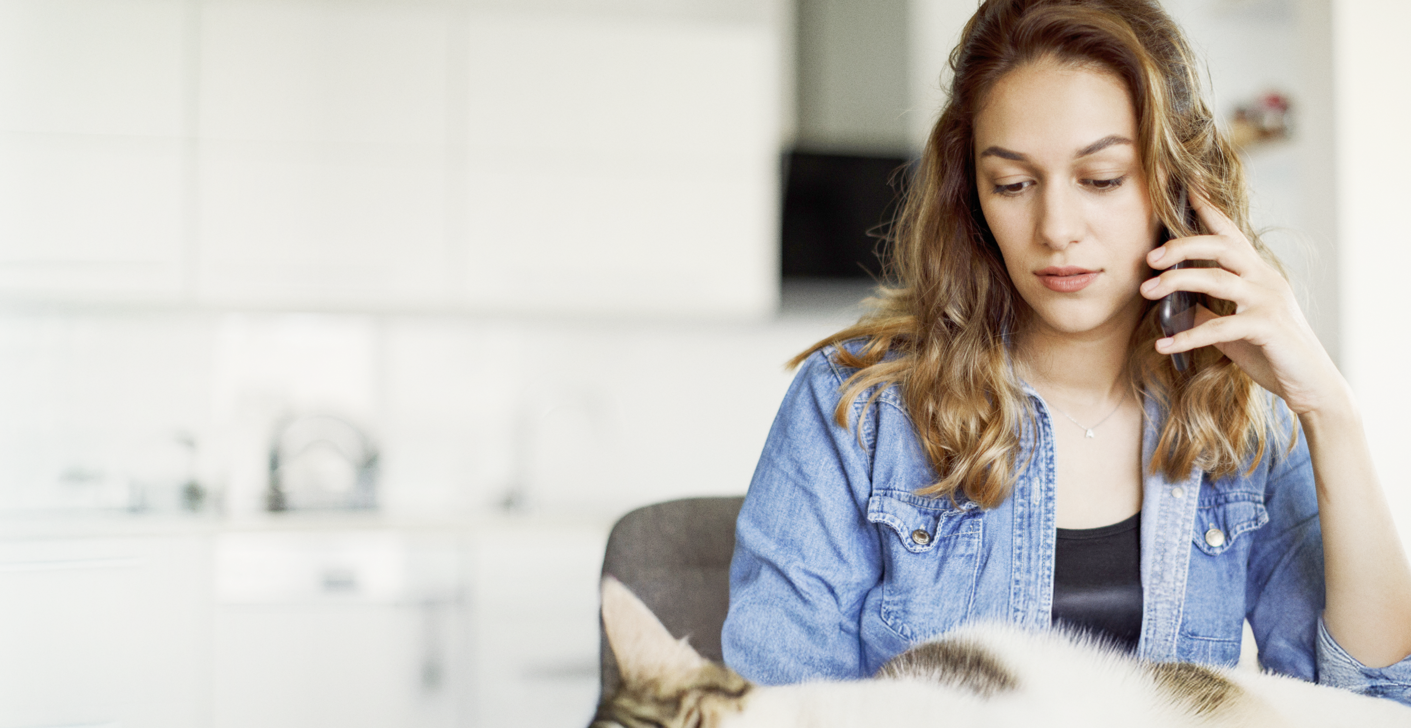 Woman in a denim shirt sitting with a cat in a bright kitchen, partially visible.